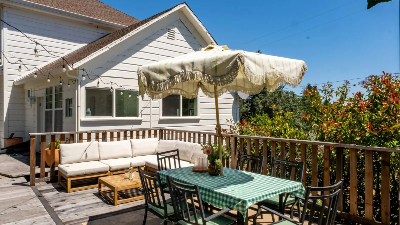 Deck dining table with gingham cloth and fringed umbrella beside the farmhouse